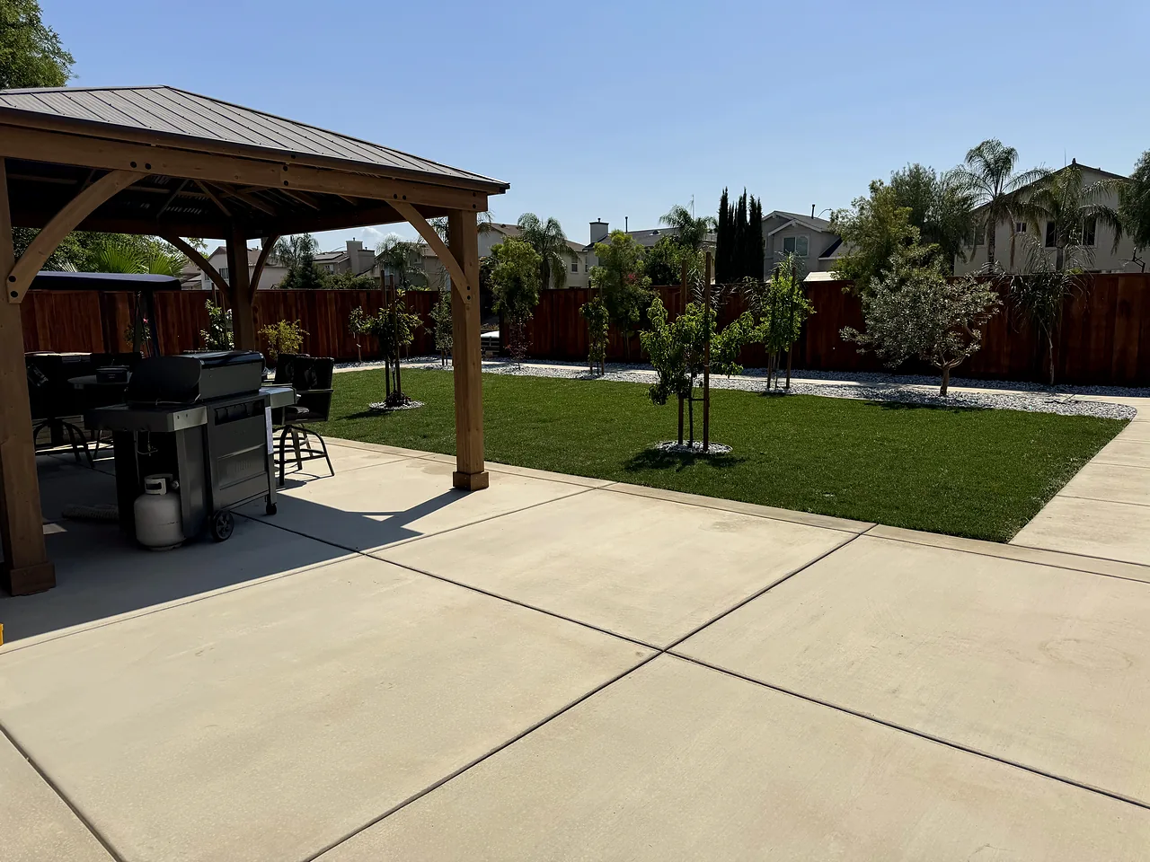 Backyard after: new concrete patio under a gazebo, fresh lawn, young trees and white rock borders